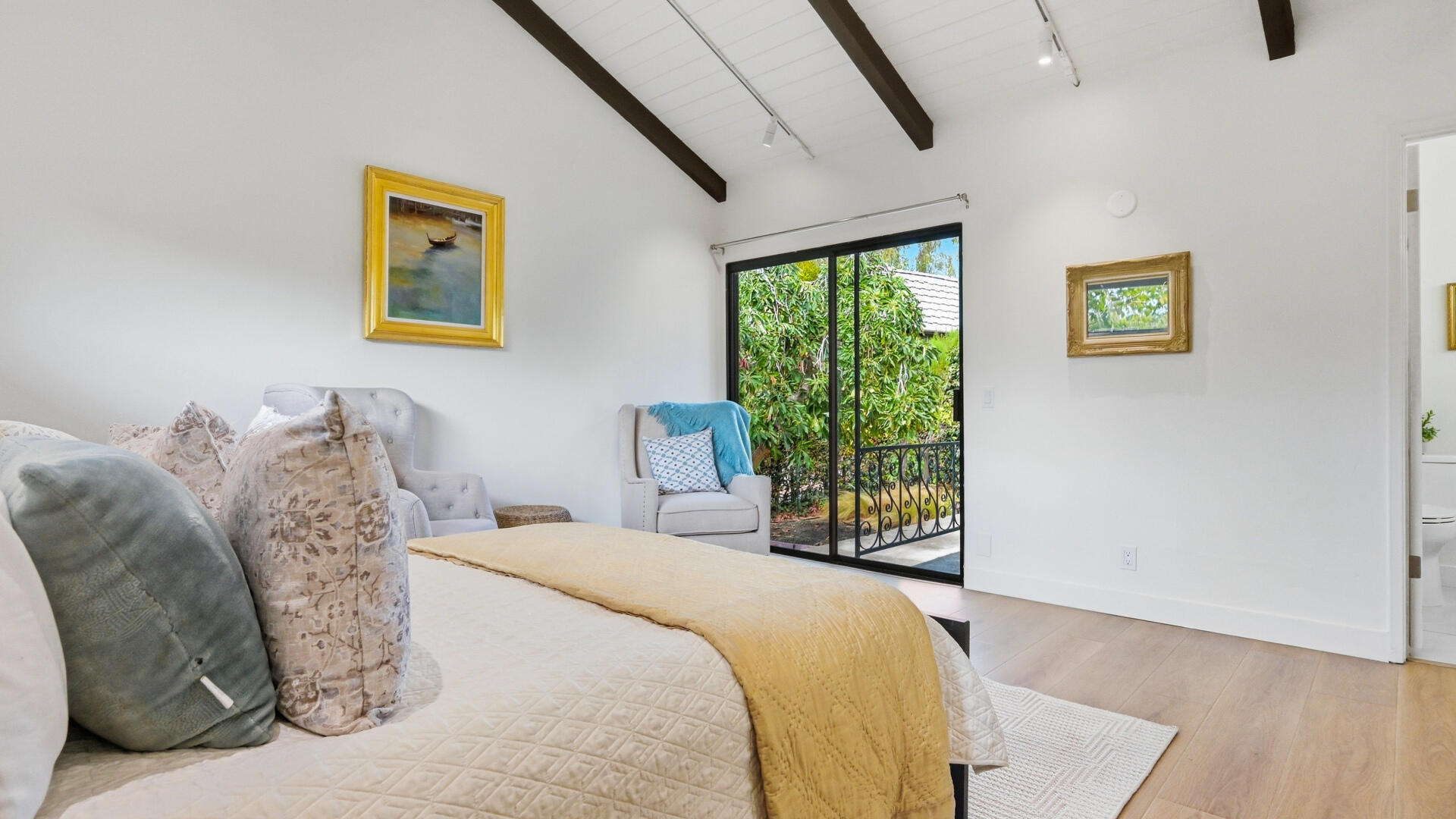 Furnished bedroom with exposed ceiling beams and sliding glass doors opening to a leafy balcony.