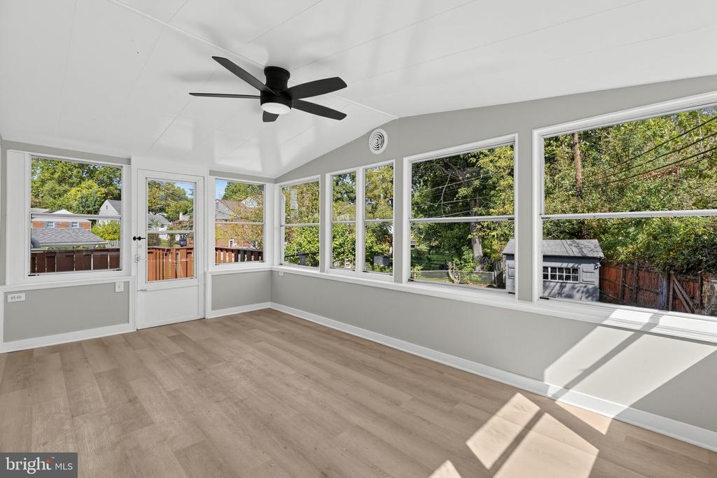 Empty sunroom with a vaulted ceiling, black fan, and large windows overlooking a tree-lined backyard.