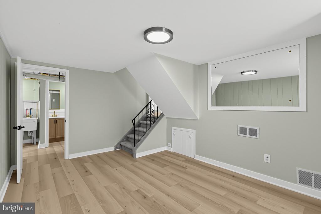 Empty basement room with light wood floors, carpeted stairs, and an open doorway to a bathroom.