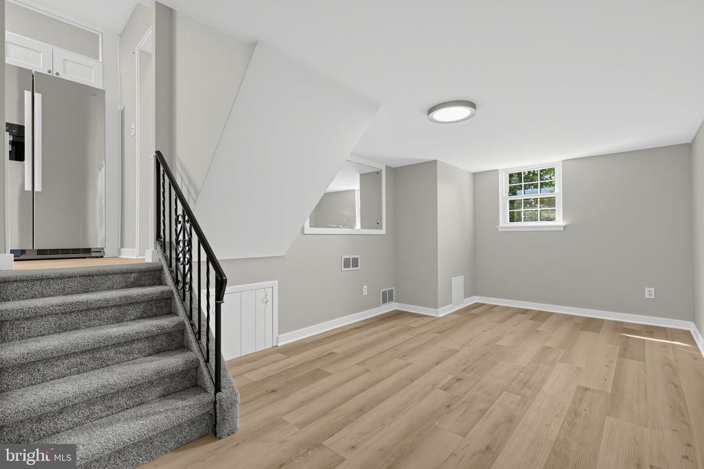 Empty renovated living area with light wood-look floors, a carpeted staircase, and stainless-steel refrigerator.