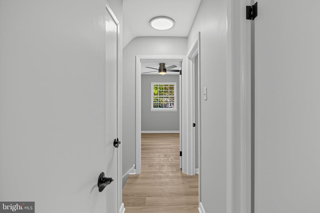 Hallway with light wood floors leading to a room with a ceiling fan and window.