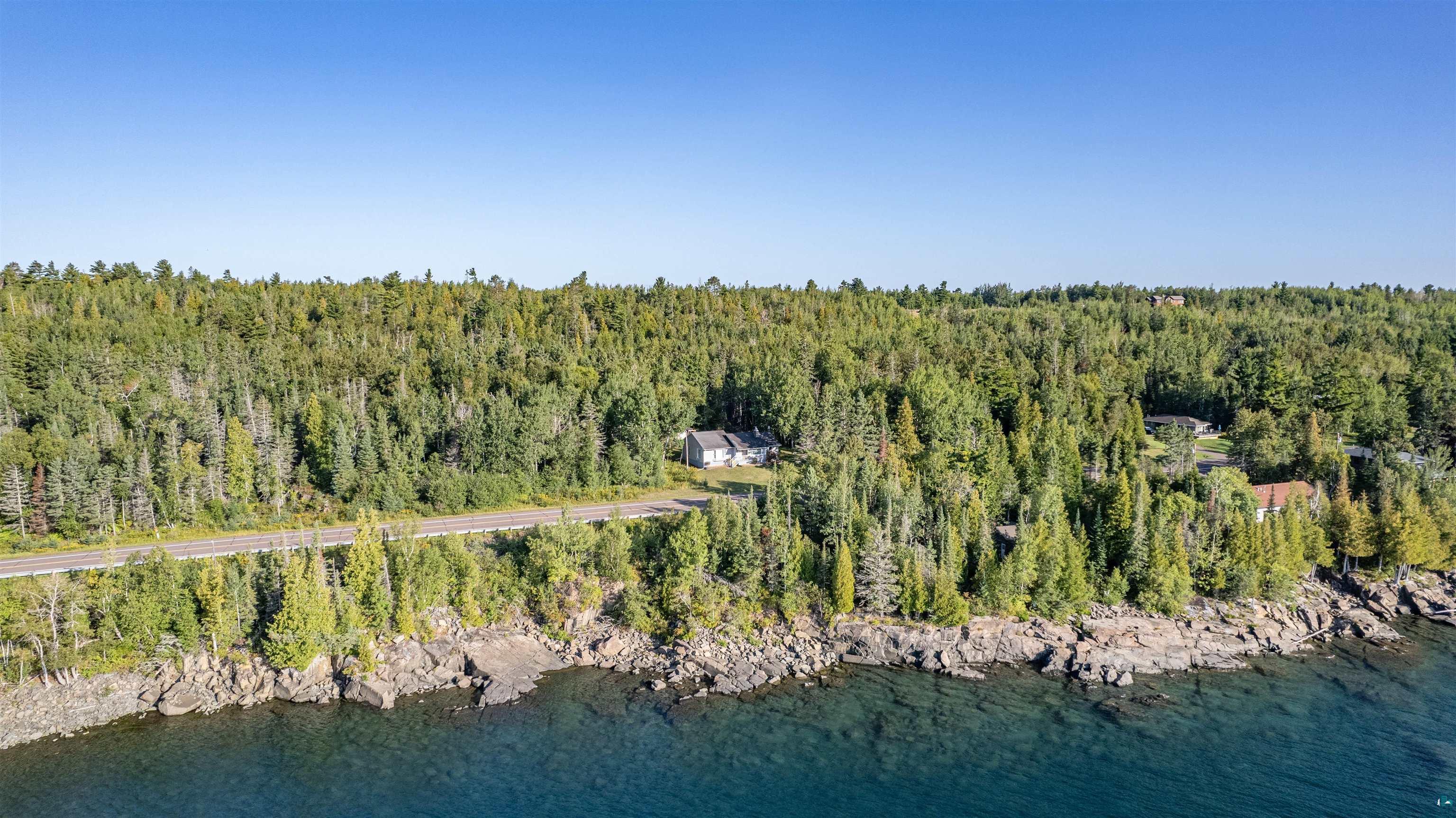 Aerial view of a forested lakeshore home beside a rocky shoreline and clear blue water.