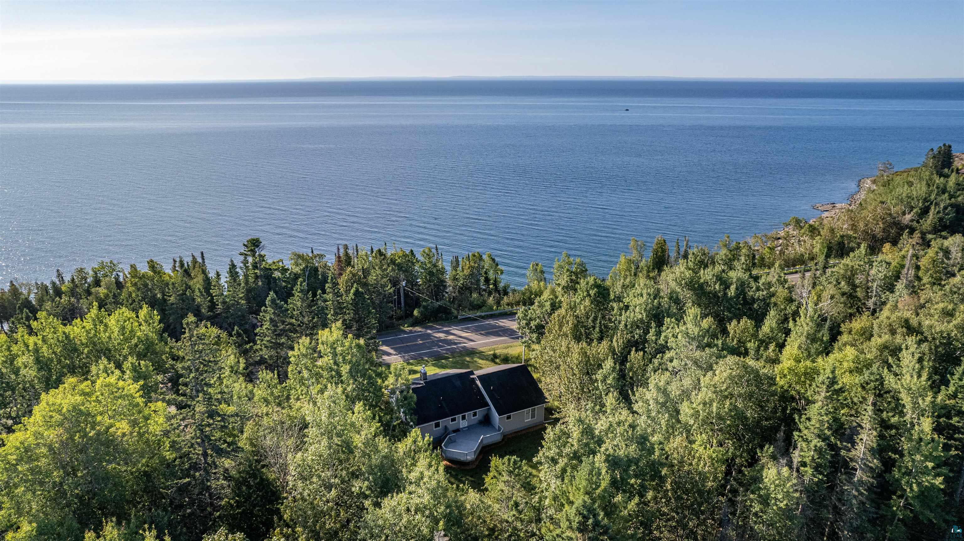 Aerial view of a wooded lakeside home with a deck, shoreline, and expansive water beyond.