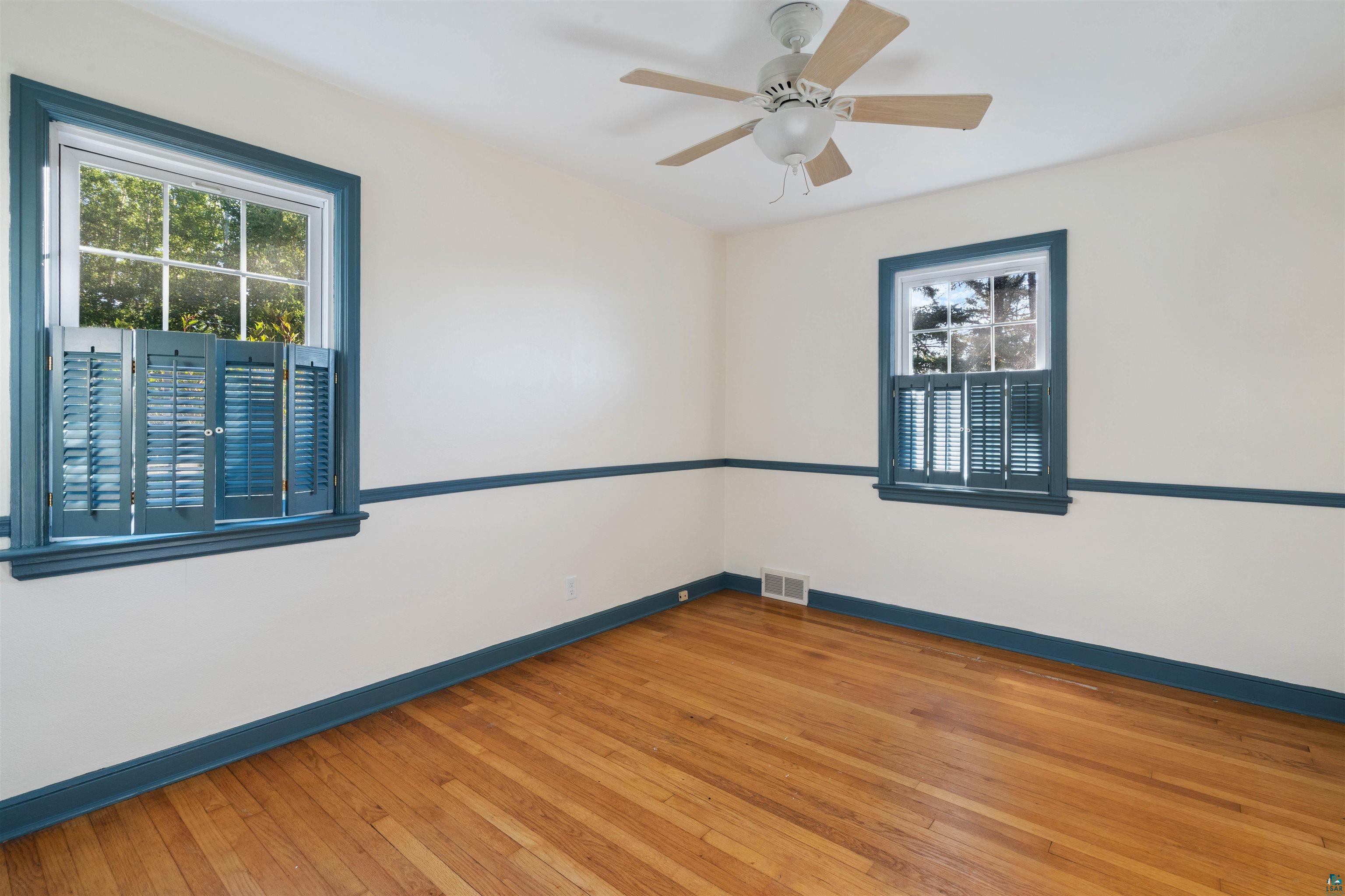 Empty room with hardwood floors, blue-trimmed windows and shutters, and a ceiling fan.