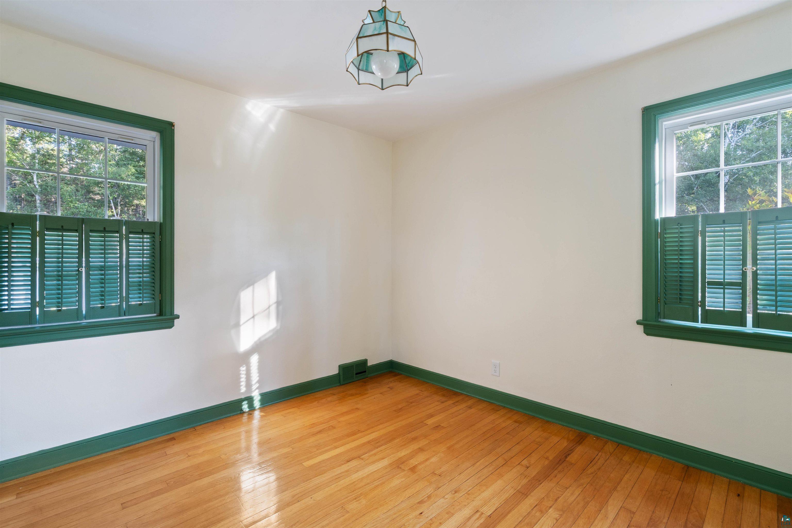Empty room with polished hardwood floors, green-trimmed shuttered windows, and a pendant light.