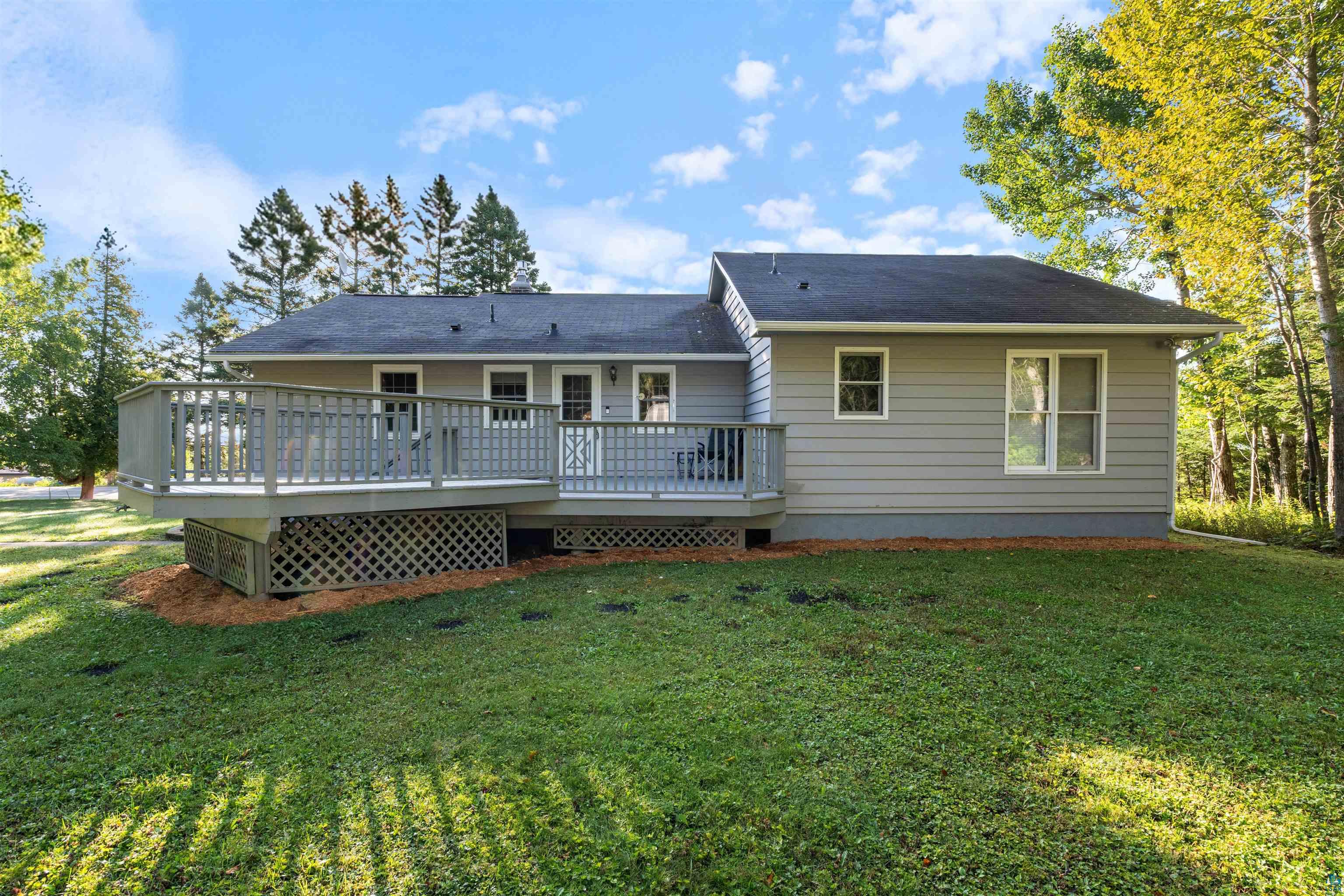 Rear of a gray-sided home with a raised deck, lawn, and surrounding trees.