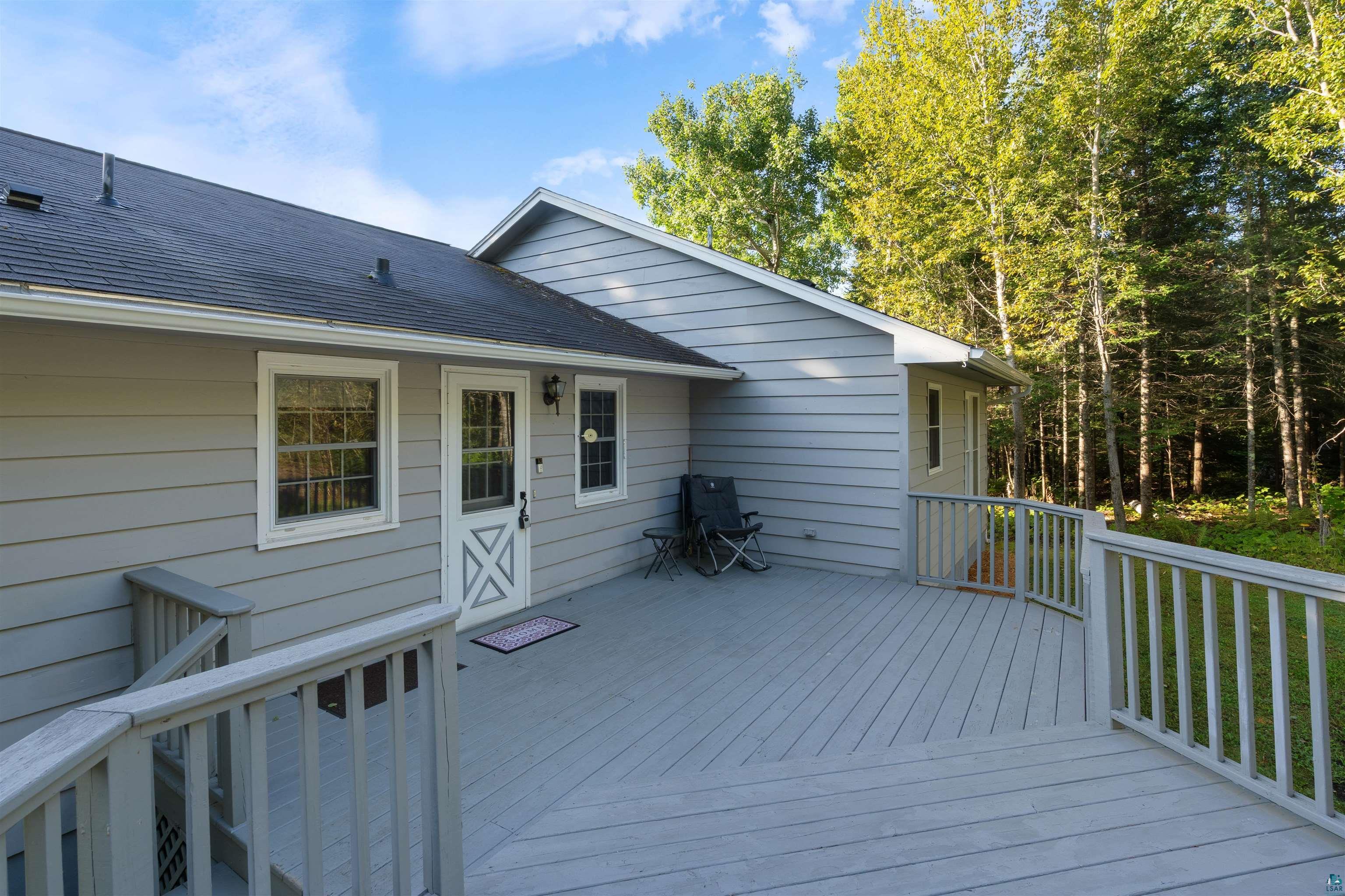 Gray wraparound deck beside a light-gray house, with white railings and woods beyond.