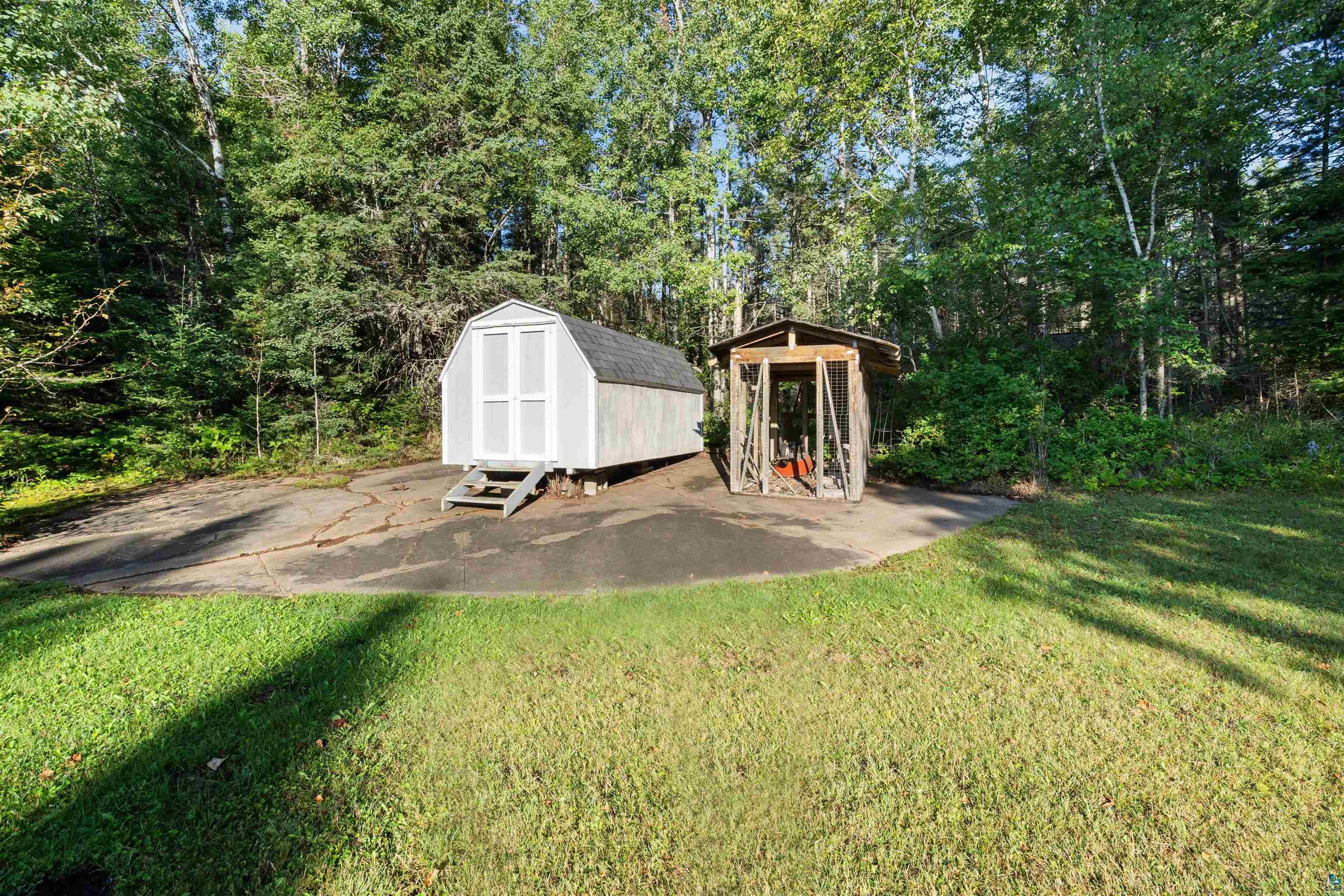 White storage shed and wooden covered enclosure on a paved clearing surrounded by trees.