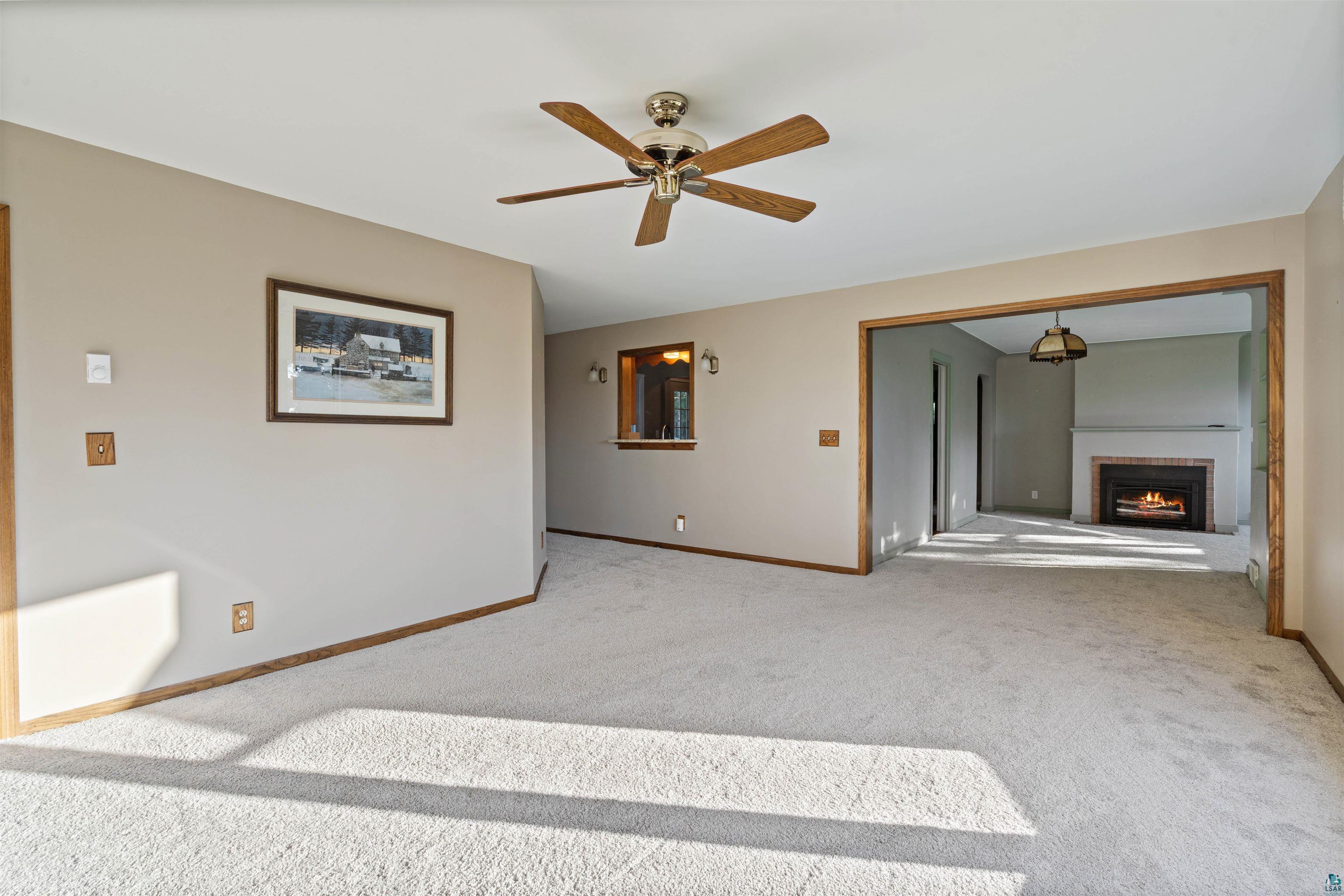 Vacant carpeted living room with a wood-blade ceiling fan and an adjoining fireplace room.