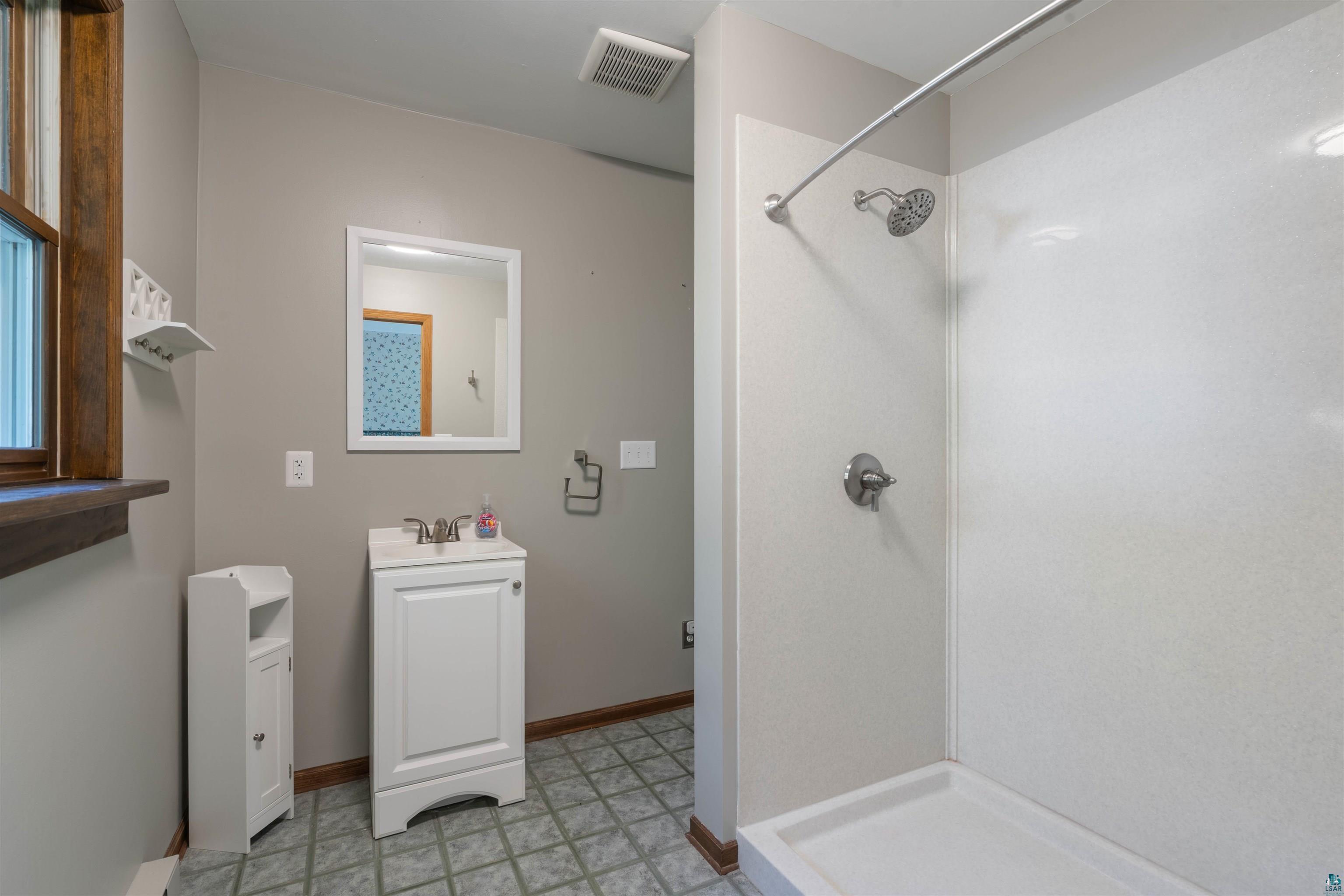 Bathroom with a white walk-in shower, compact vanity, gray tile floor, and wood-framed window.