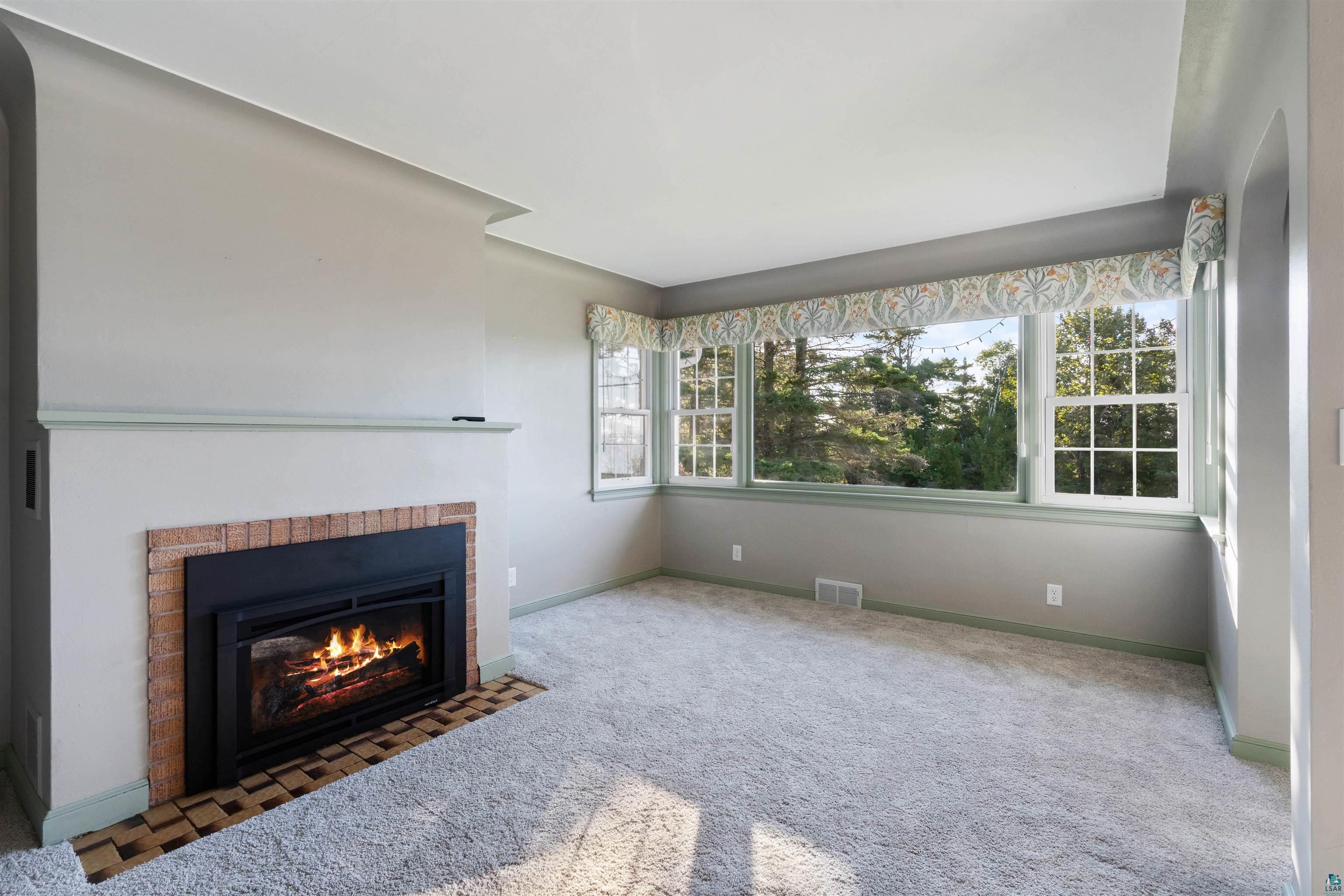 Vacant living room with a brick fireplace and wide windows overlooking trees.
