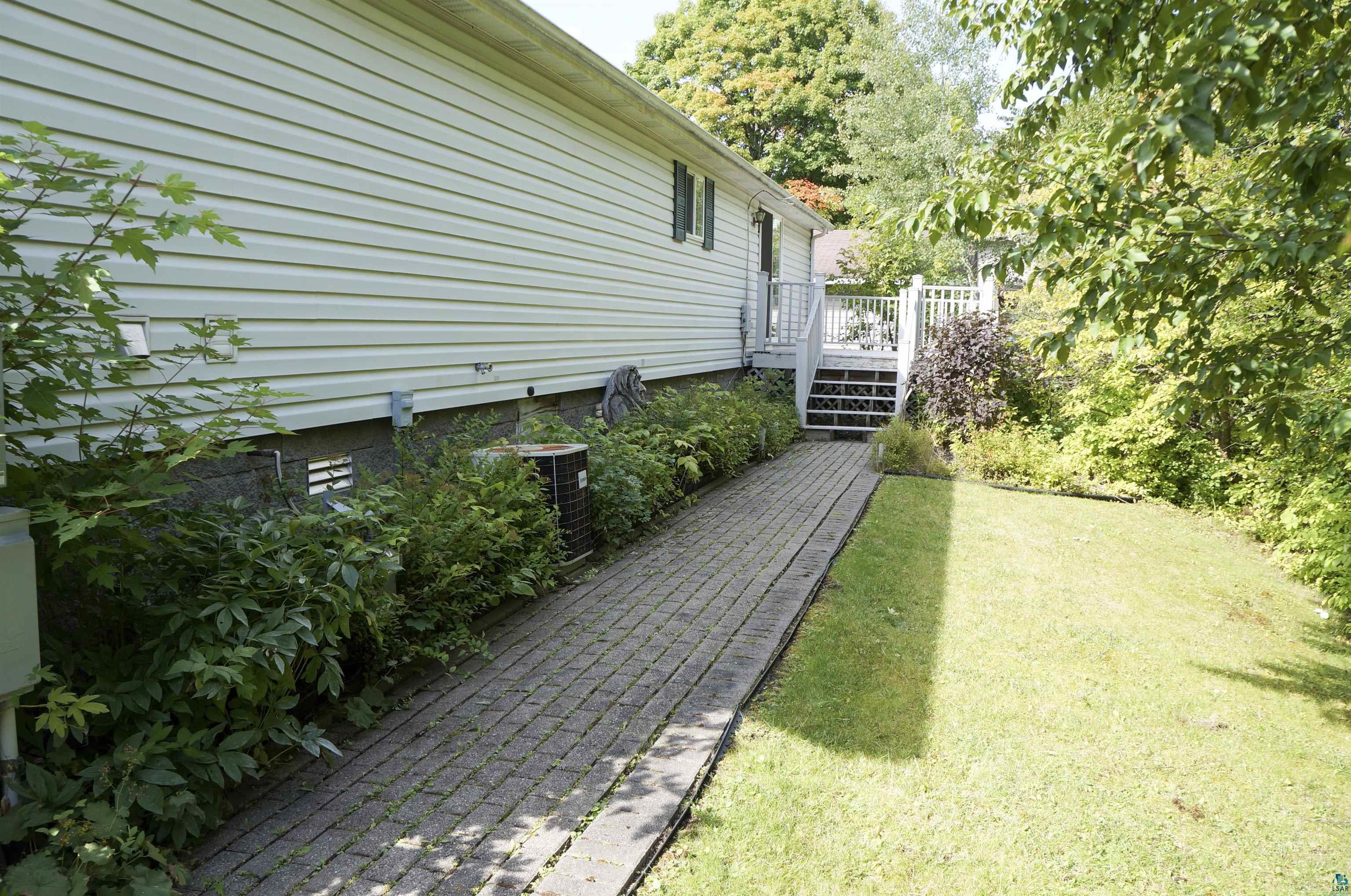 House side yard with a brick walkway, raised deck, lawn, and mature trees.