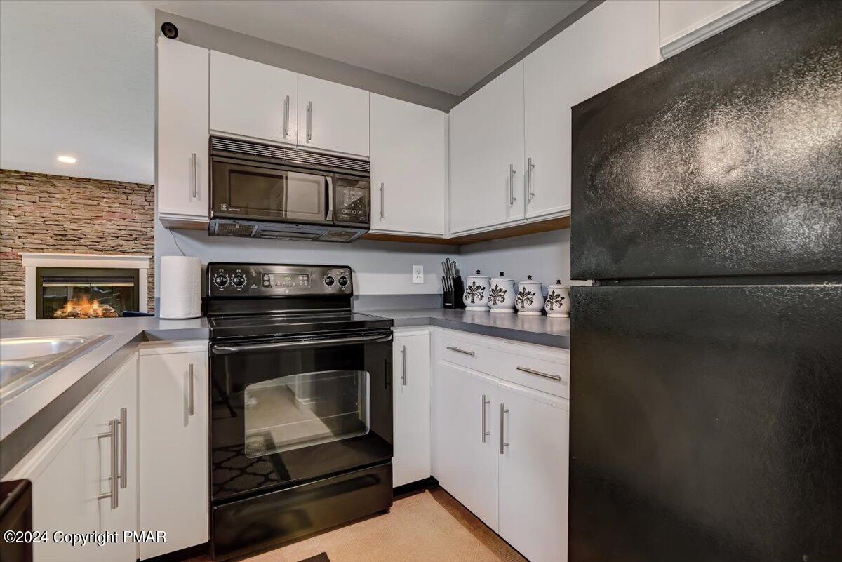 Kitchen with white cabinets and black appliances, opening toward a stone fireplace.