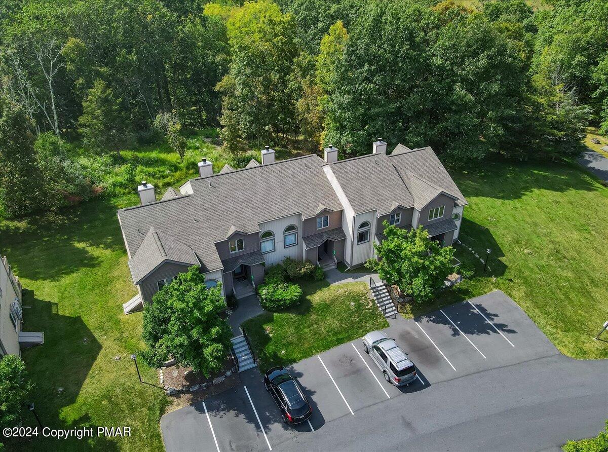 Aerial view of attached homes beside a parking lot, lawn, and wooded grounds.