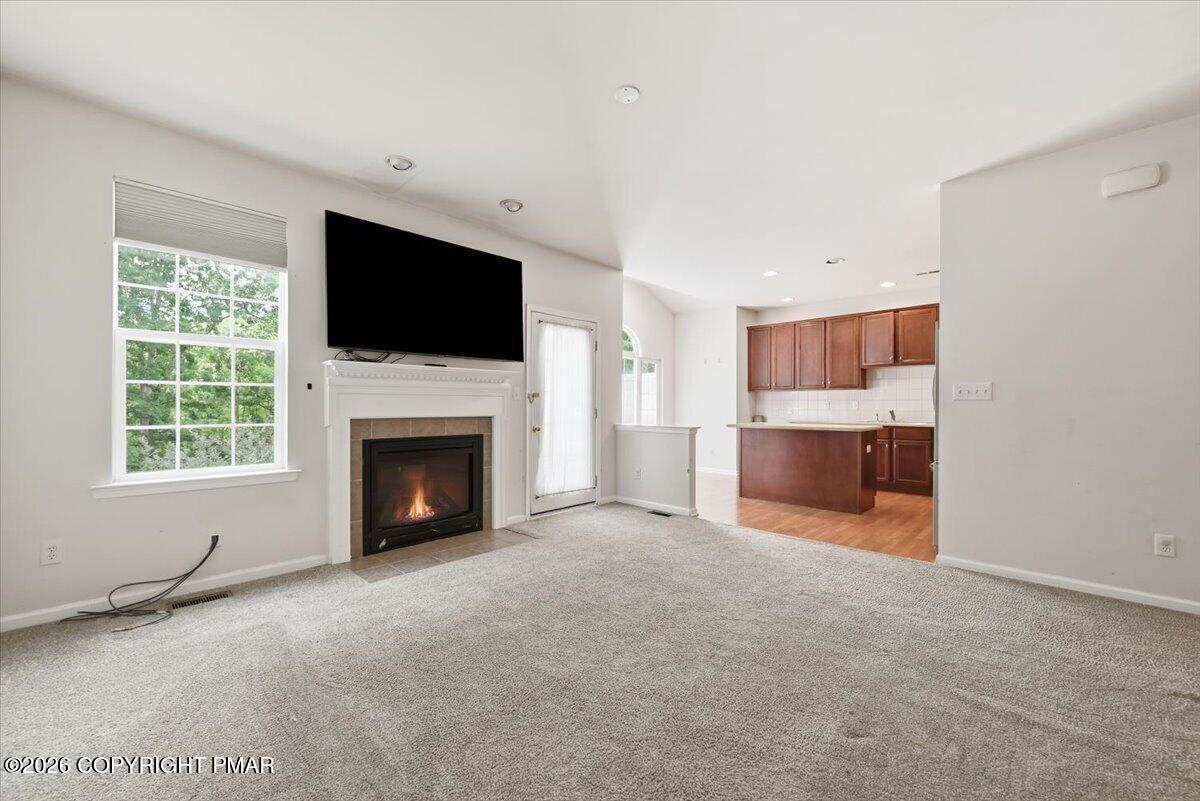 Vacant living room with a lit fireplace, wall-mounted TV, beige carpet, and adjoining wood-cabinet kitchen.