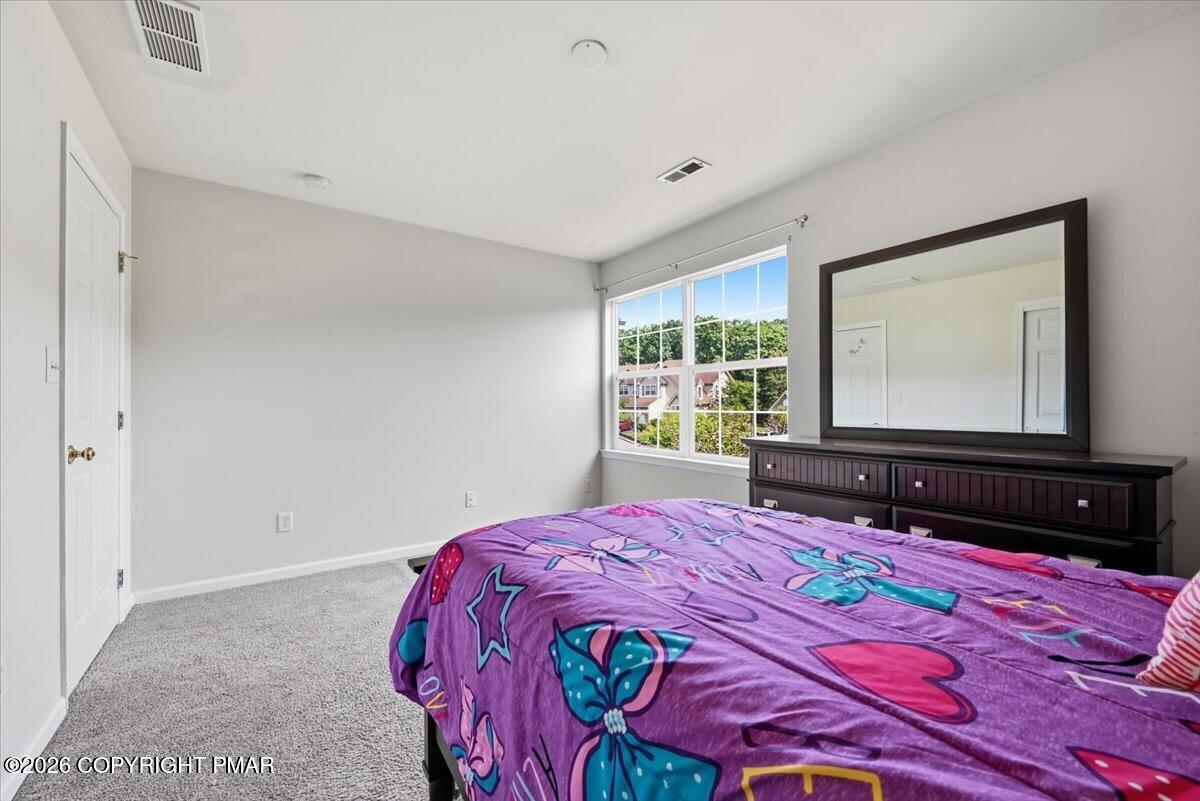 Bedroom with a colorful purple bedspread, gray carpet, and a large window overlooking trees and houses.