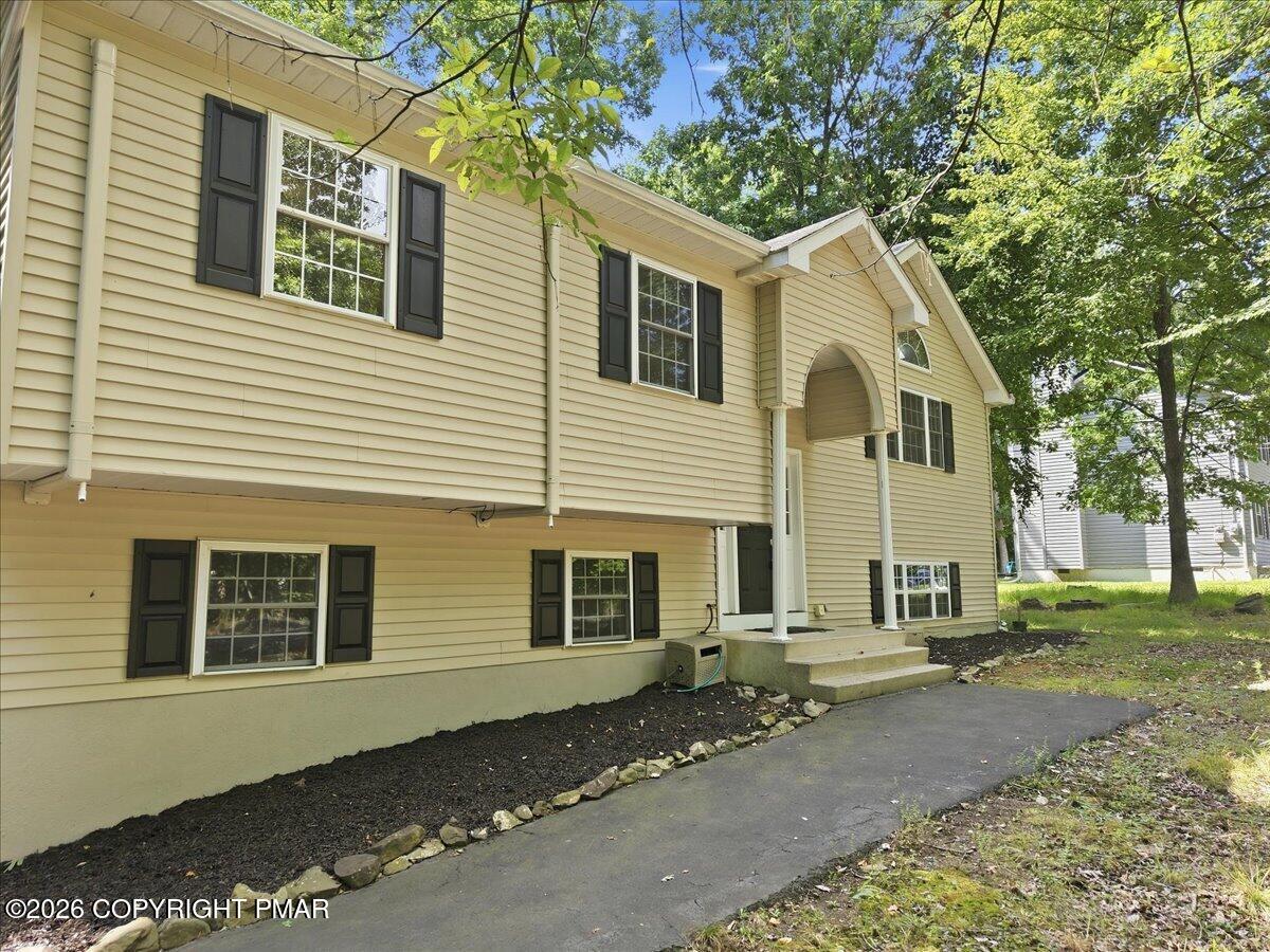 Beige two-story home with dark shutters, a covered entry, and a paved walkway beneath leafy trees.