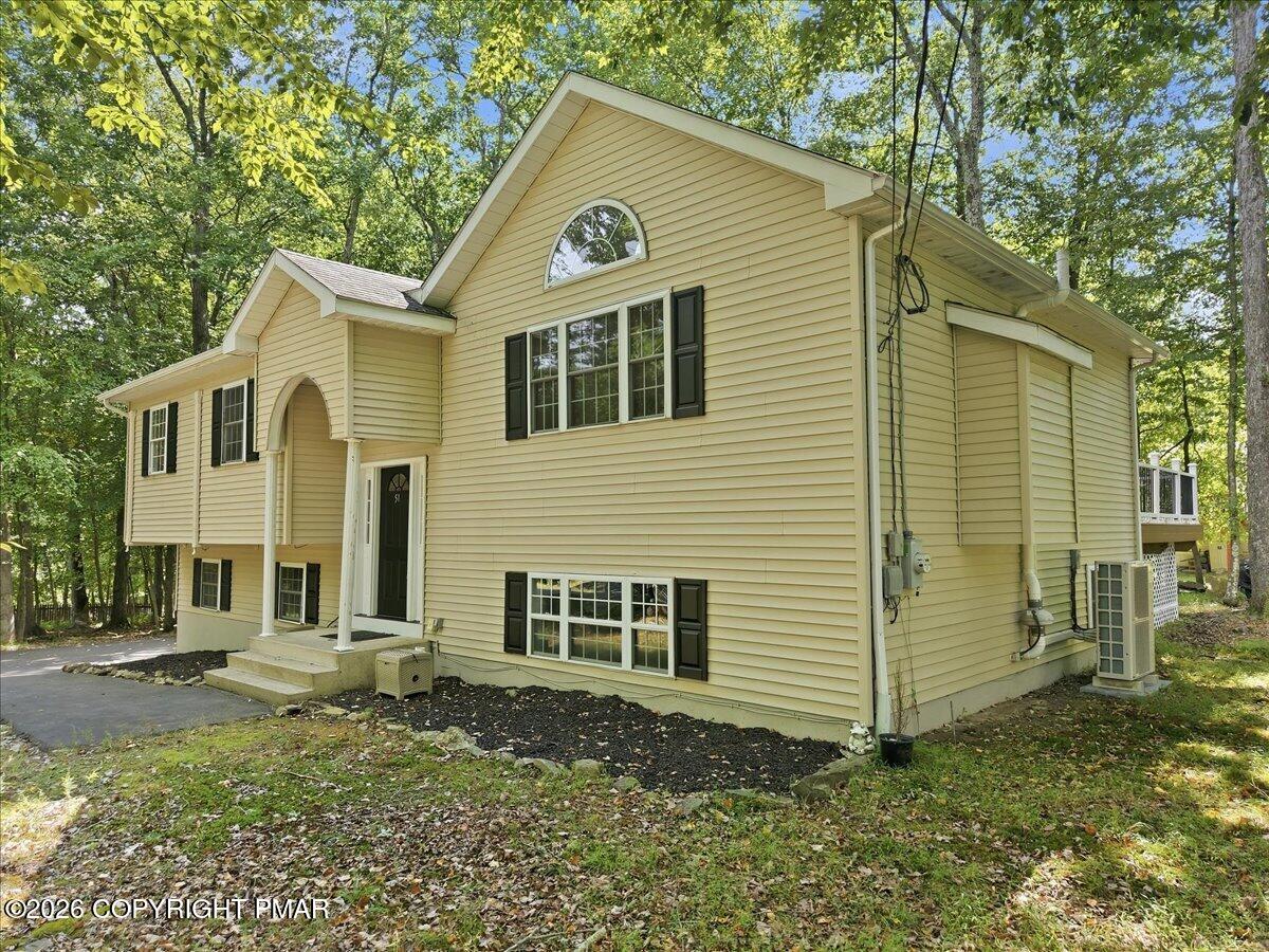 Pale yellow split-level house with dark shutters and an arched entry, surrounded by mature trees.