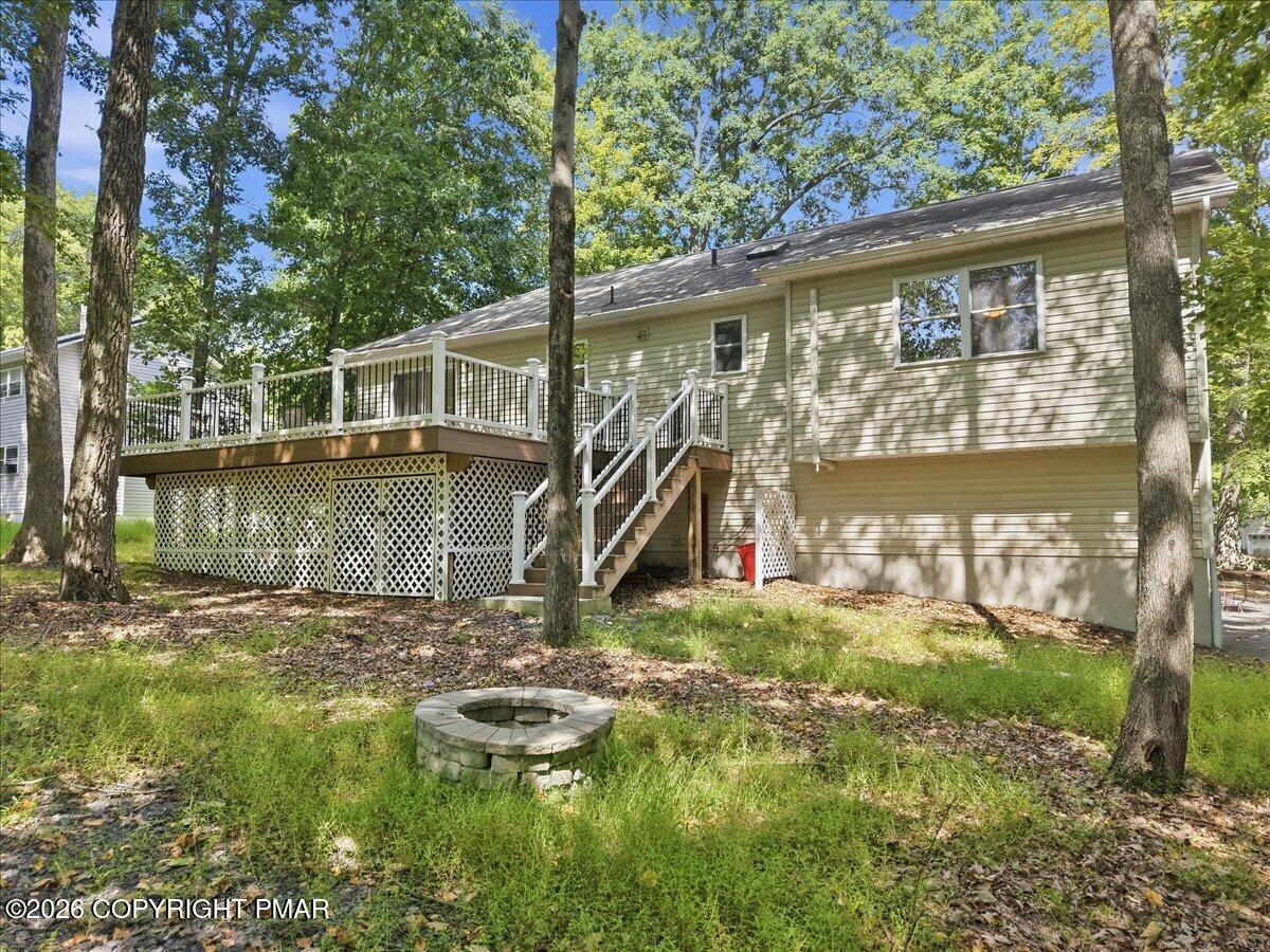 Rear of a light-colored house with a raised deck and stone fire pit in a wooded yard.