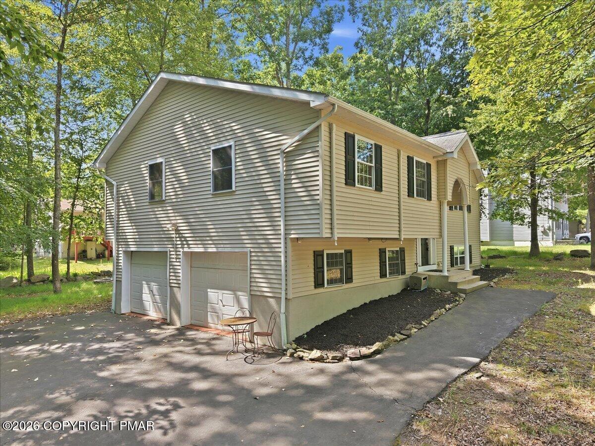 Beige split-level home with two garage doors and a paved driveway beneath mature trees.
