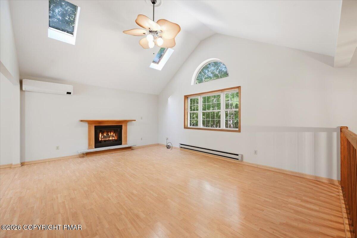 Empty living room with a lit fireplace, vaulted ceiling, skylights, and light hardwood floors.