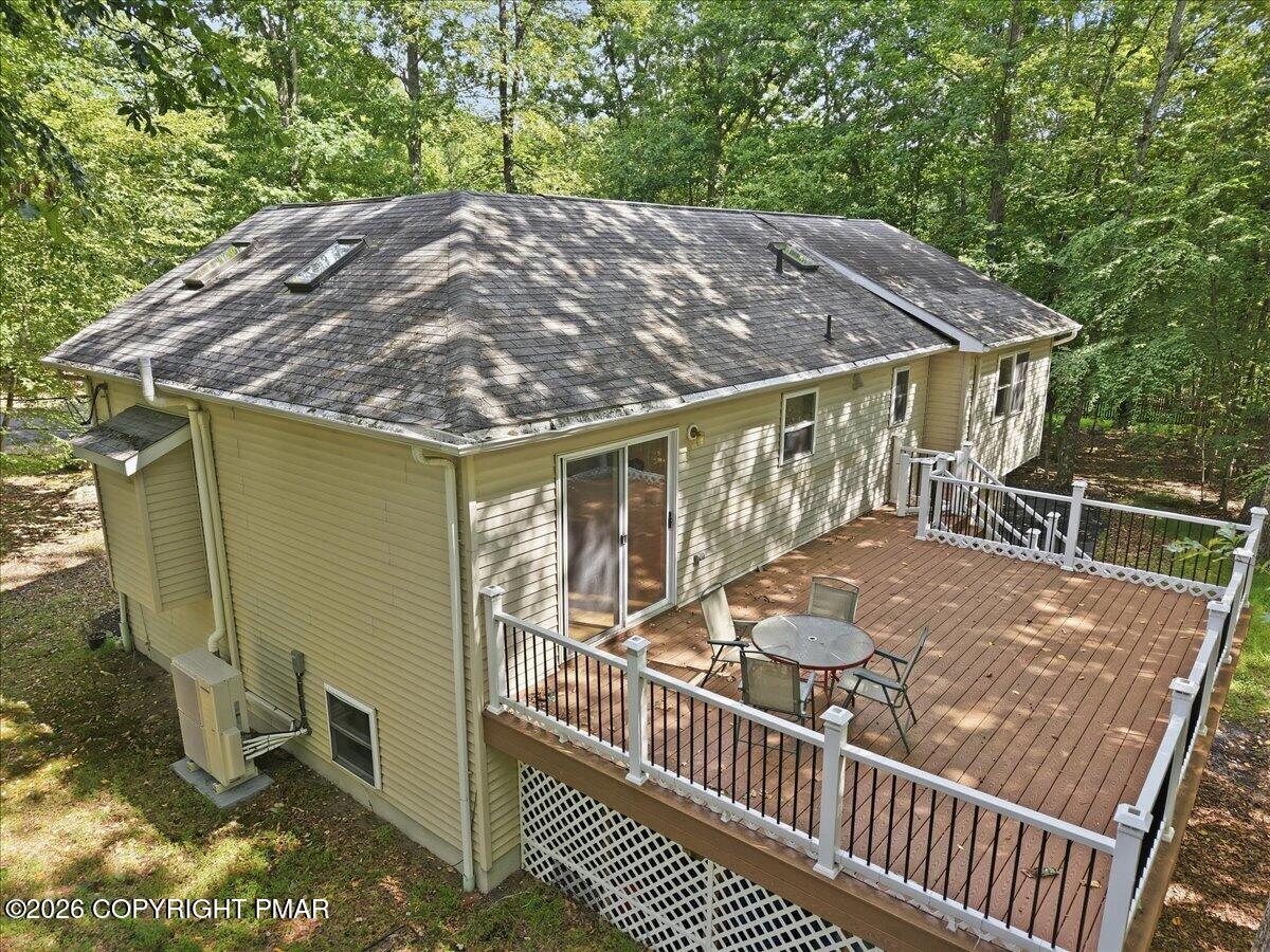 Rear of a beige house with a large wooden deck and patio furniture, surrounded by leafy trees.