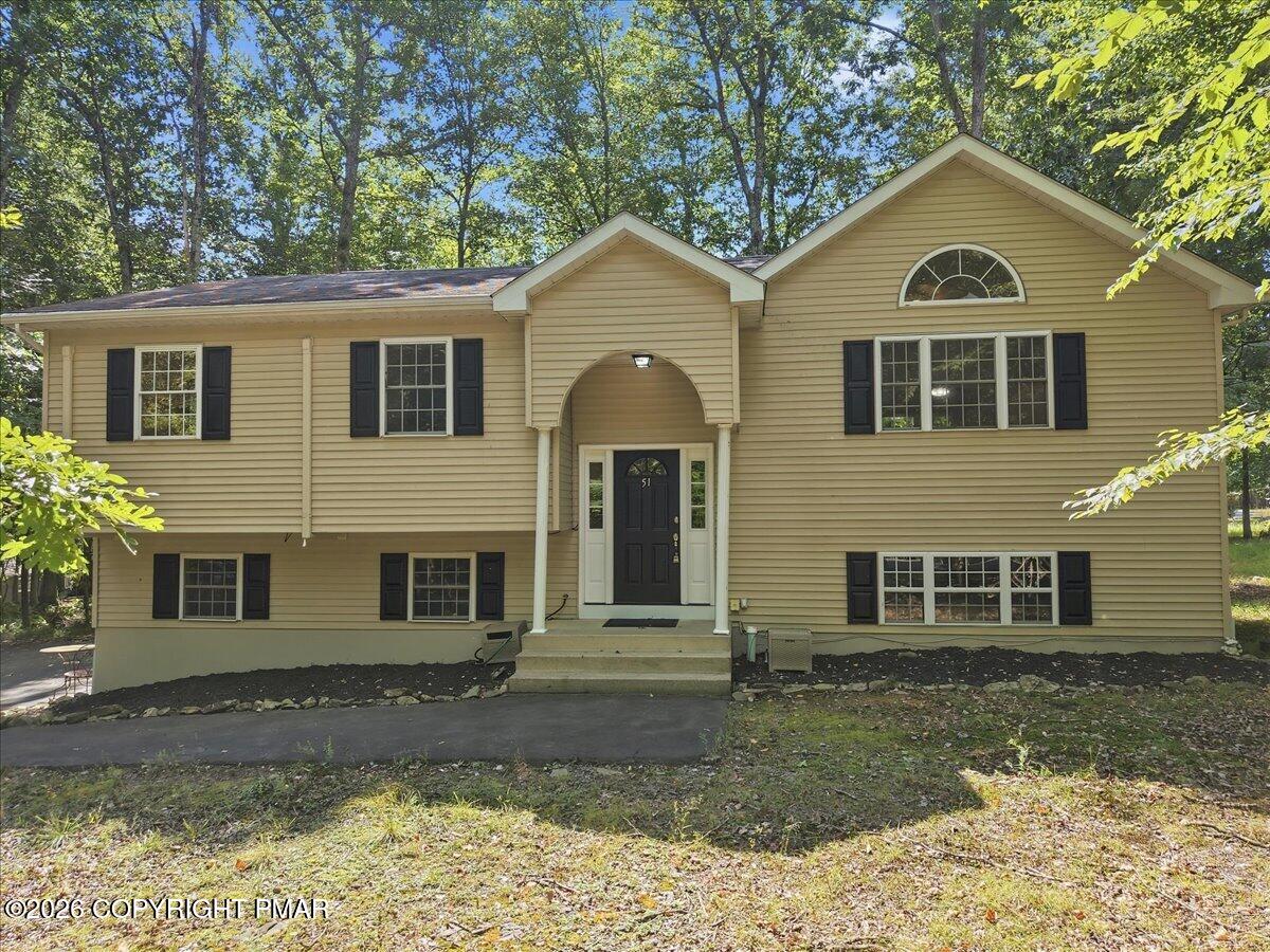 Tan split-level house with black shutters, an arched entry, and leafy trees surrounding it.