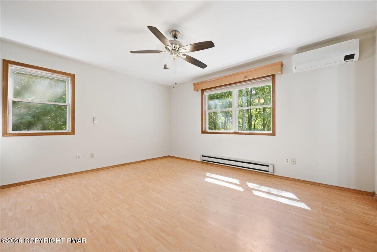 Empty room with light wood flooring, two windows, a ceiling fan, and wall-mounted air conditioner.