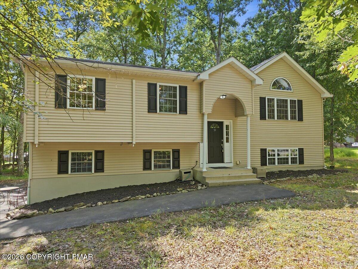 Beige split-level home with black shutters and a covered arched entry among mature trees.