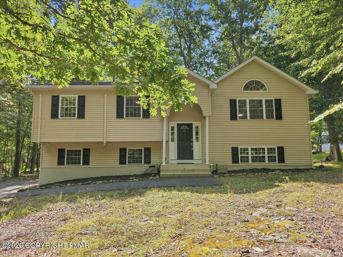 Beige split-level home with black shutters and a covered entry beneath leafy trees.