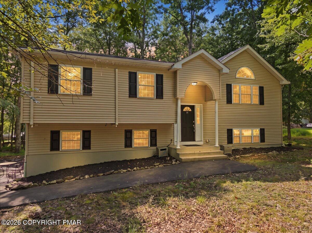 Beige split-level home with black shutters and an arched covered entrance beneath mature trees.
