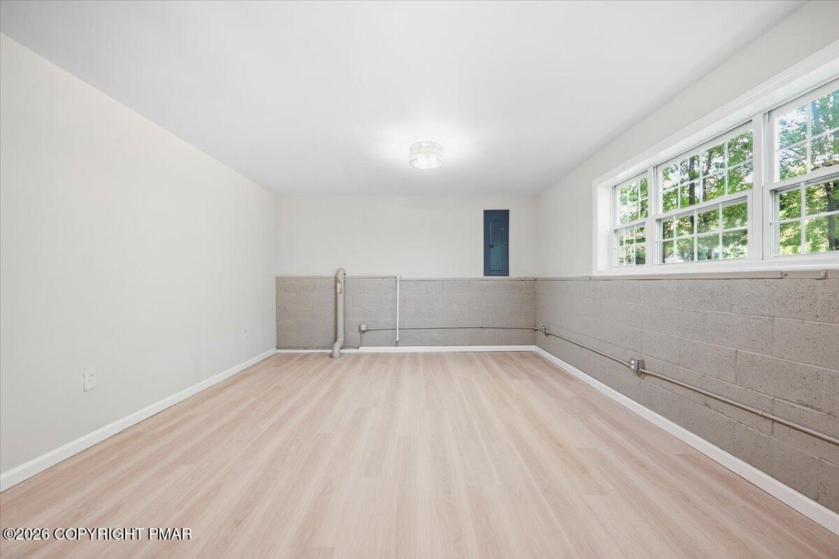 Vacant basement room with pale wood-look flooring, exposed block walls, utility pipes, and large windows.