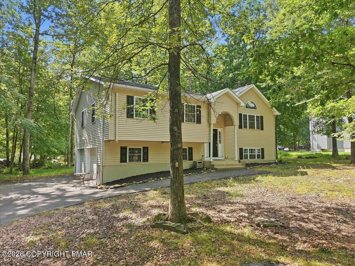 Yellow split-level home with an arched entry and dark shutters among mature trees.