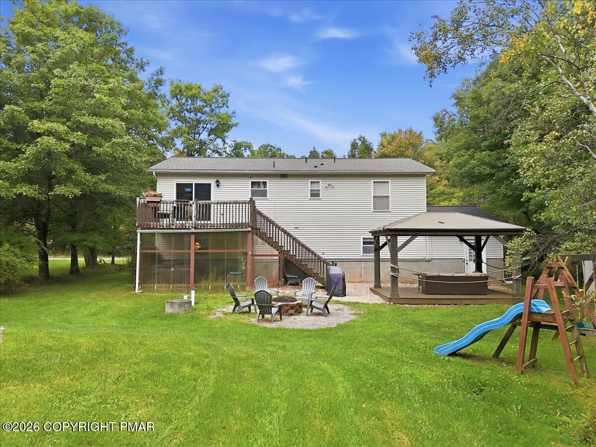 White-sided house behind a lawn, with a raised deck, gazebo, fire-pit seating area, and playset.
