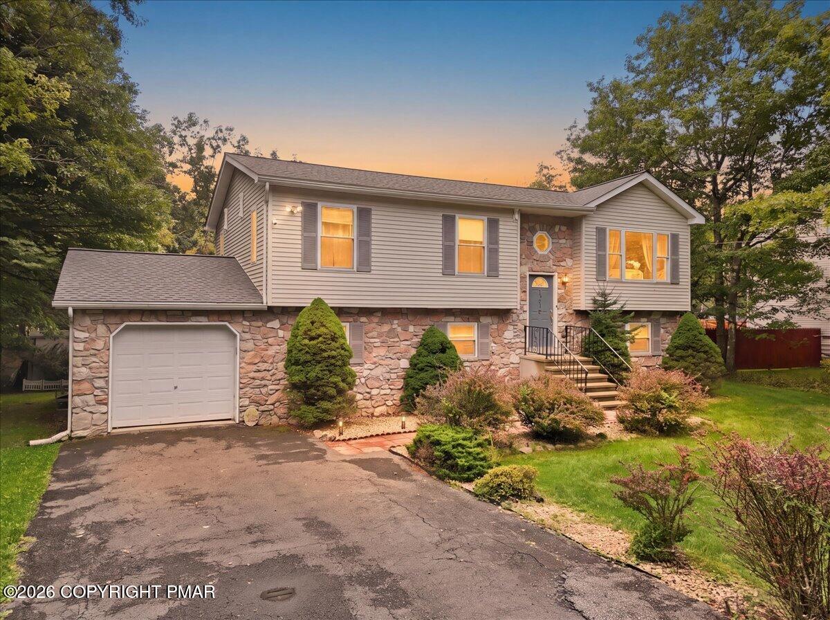 Split-level home with stone facade, attached garage, and landscaped front yard at dusk.