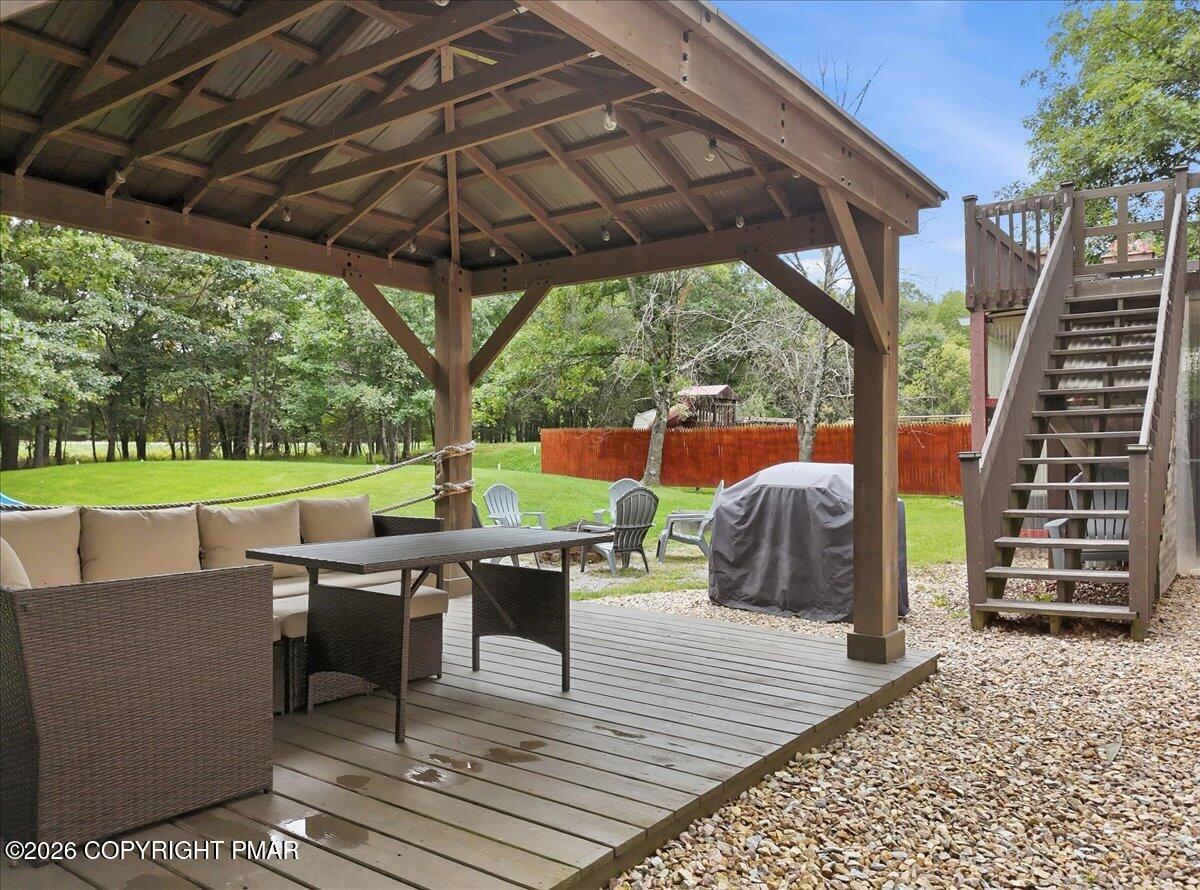 Covered wooden deck with wicker seating, dining table, and stairs beside a grassy yard.