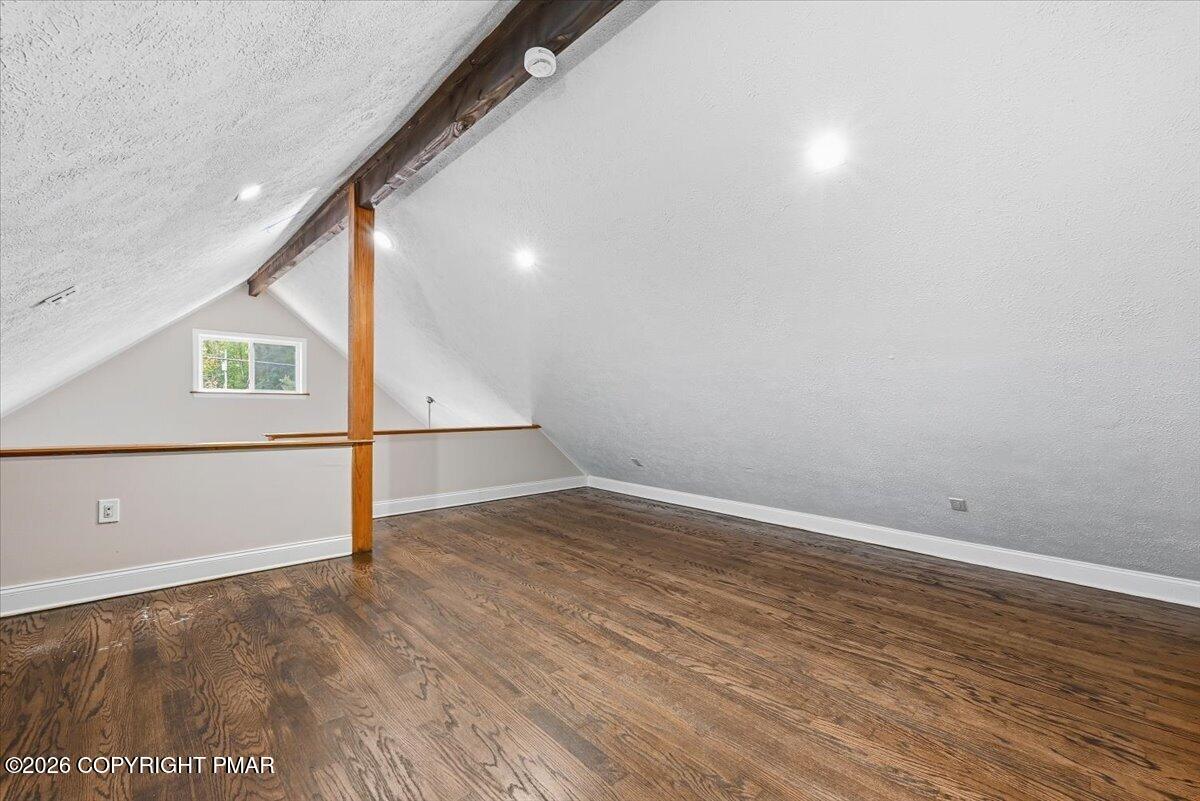 Finished attic loft with sloped ceilings, exposed wood beams, a small window, and hardwood floors.