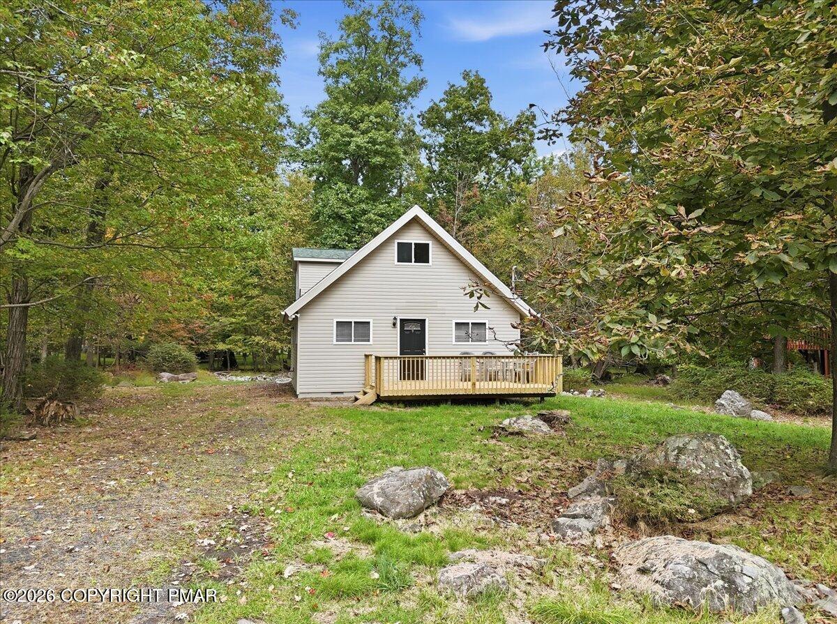 Light-sided A-frame home with a raised wooden deck, set among mature trees and rocks.