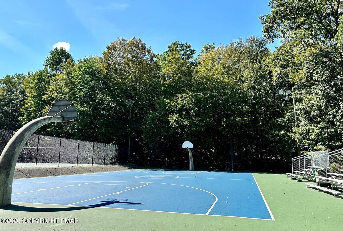 Outdoor basketball court with two hoops and bleachers beneath mature trees.