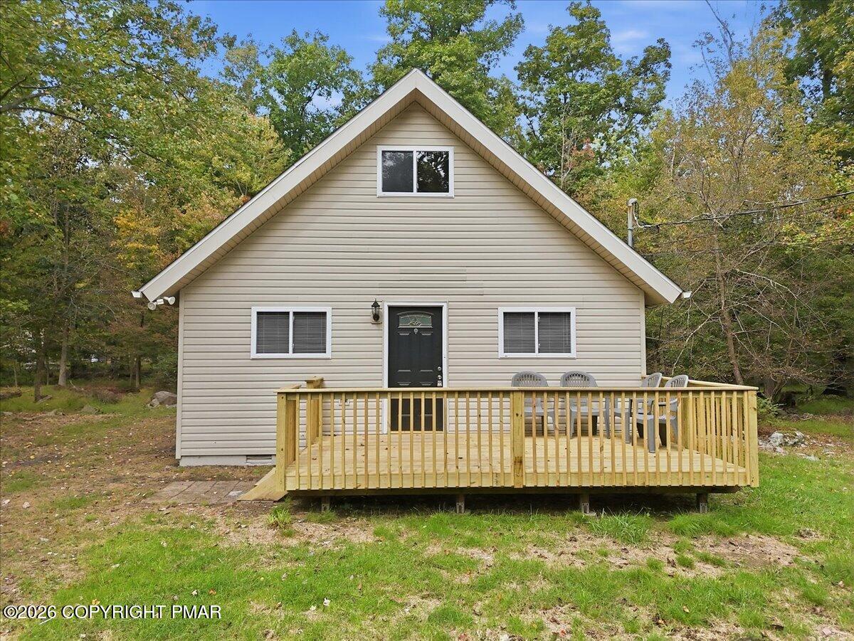 Beige gabled home with a raised wooden deck and patio chairs, surrounded by trees.
