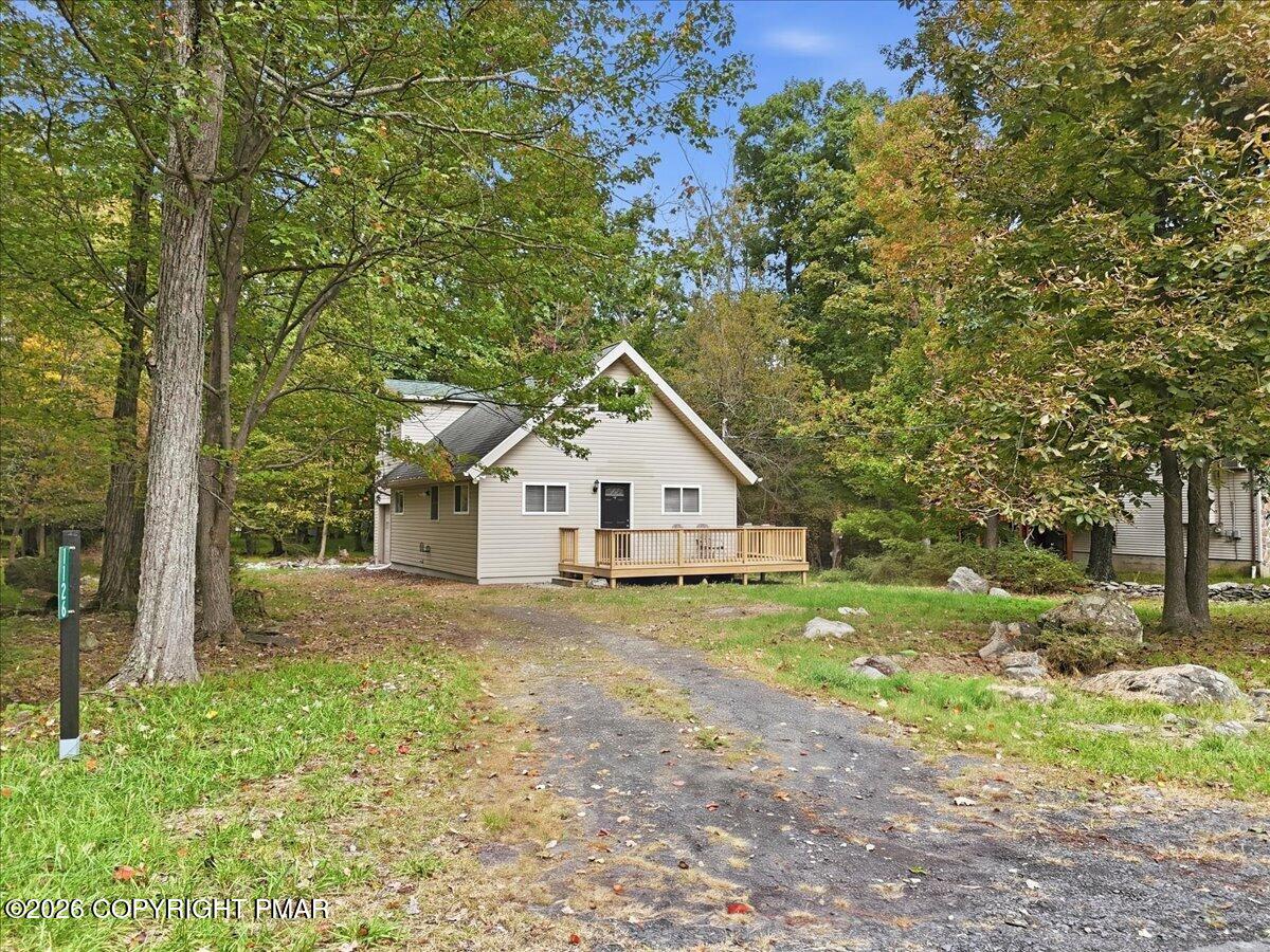Beige gabled house with a wooden porch, surrounded by autumn trees at the end of a paved driveway.