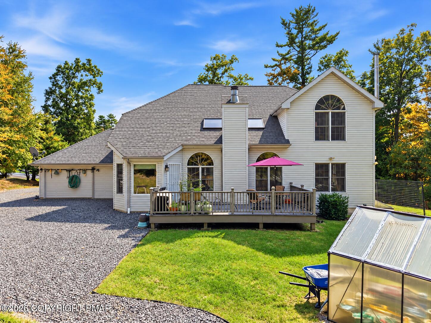 House rear exterior with raised deck, pink patio umbrella, gravel driveway, and greenhouse.