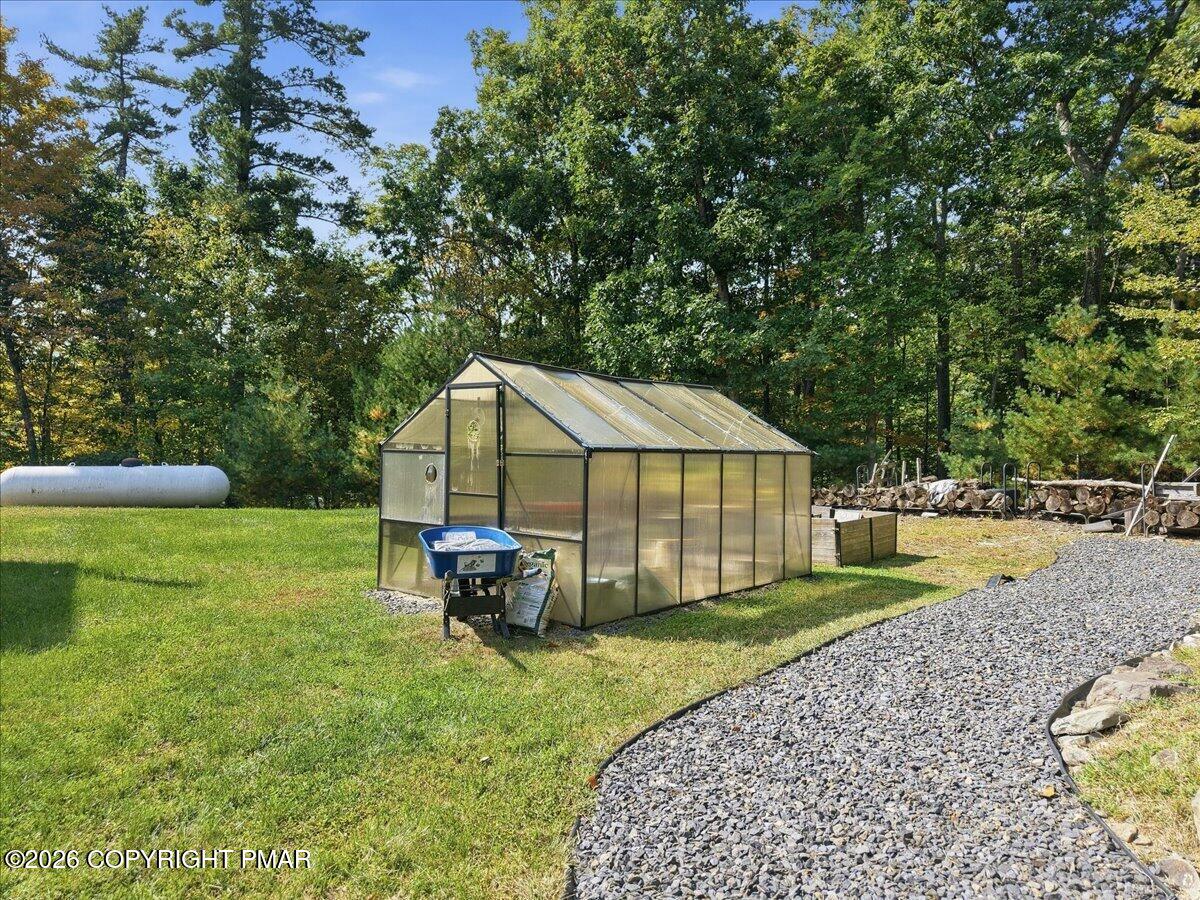 Greenhouse in a wooded yard beside a gravel path and stacked firewood.