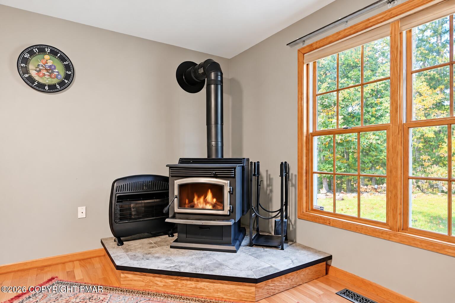 Wood-burning stove with a fire on a raised tiled hearth beside a large wood-trimmed window.