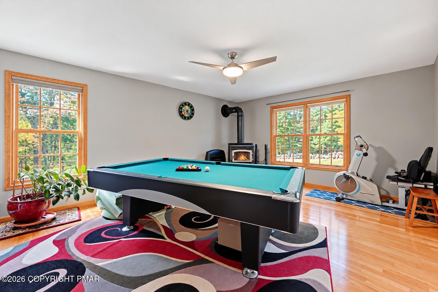 Game room with a pool table, wood-burning stove, exercise equipment, and large wood-trimmed windows.