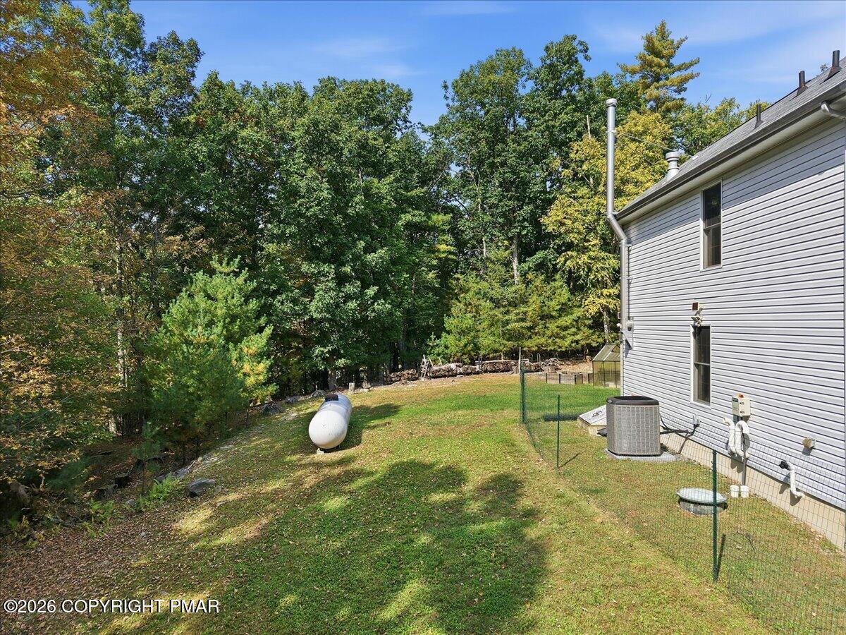 Fenced side yard beside a gray-sided home, with a propane tank and wooded border.
