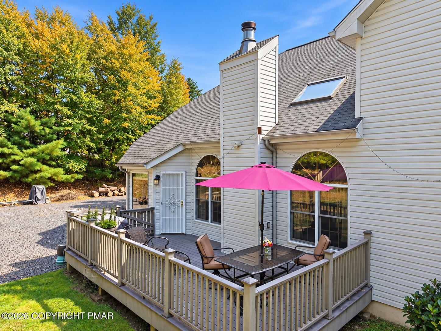 Home’s rear exterior with a furnished raised deck, pink umbrella, arched windows, and autumn foliage.