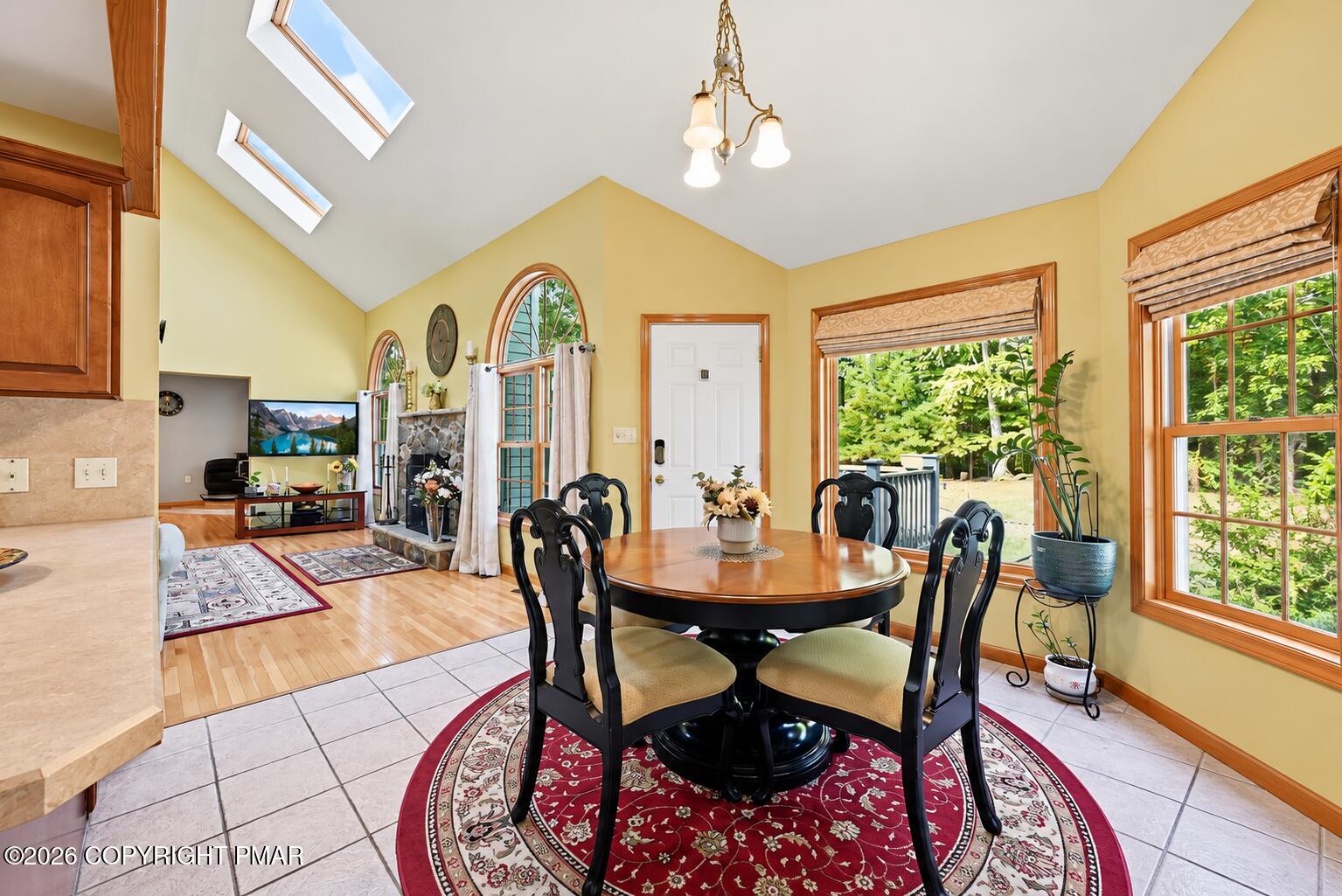 Dining area with a round wooden table, black chairs, skylights, and an adjoining living room with a stone fireplace.
