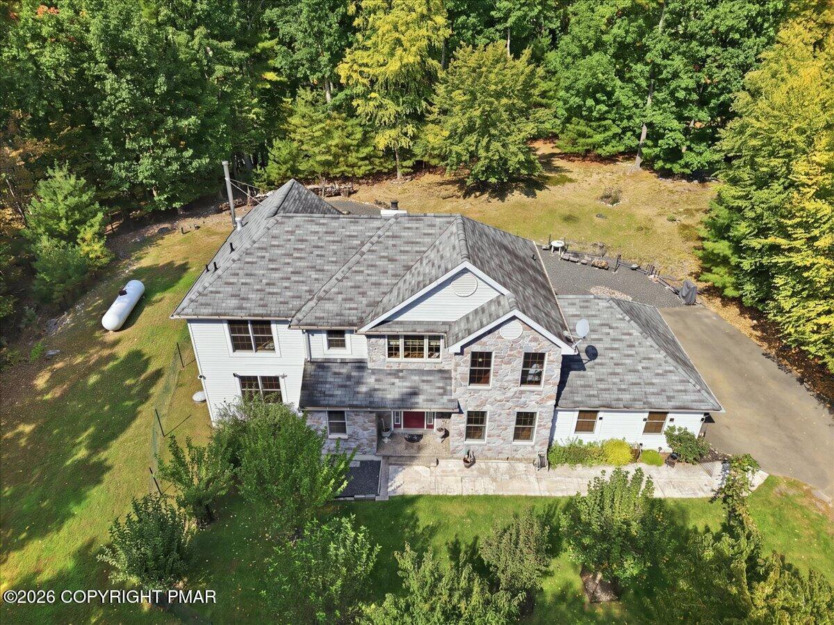 Aerial view of a stone-and-siding home with a broad shingle roof, patio, and wooded yard.