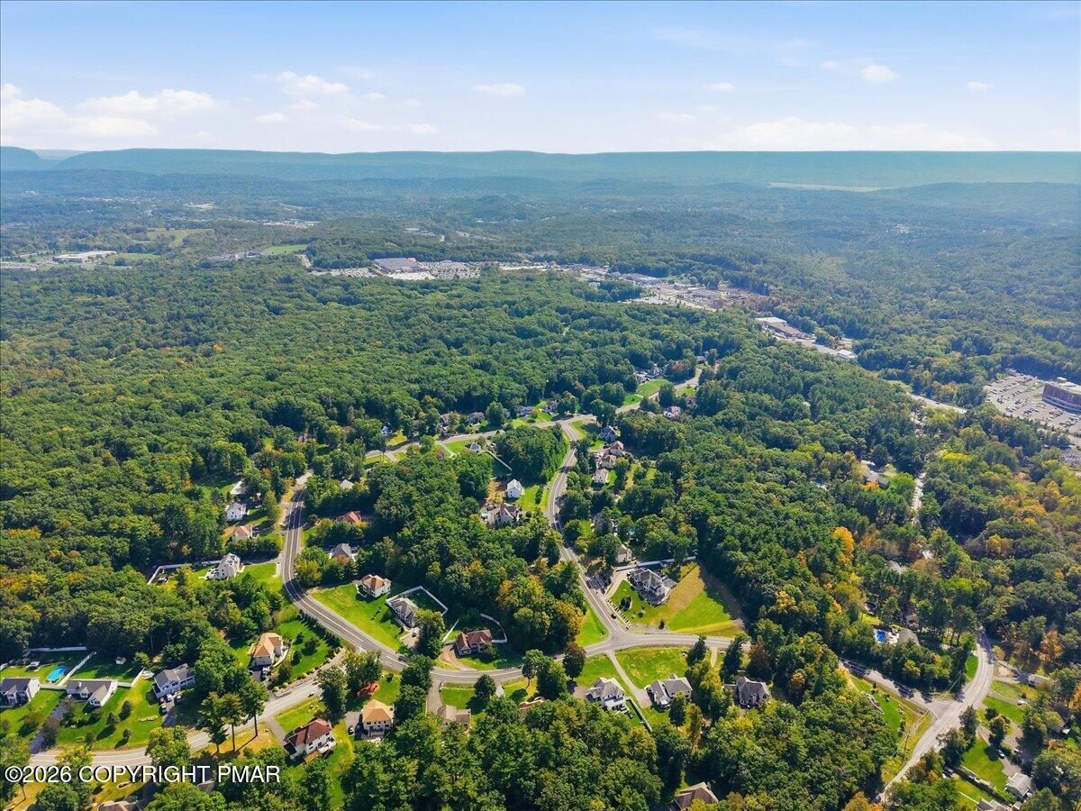 Aerial view of homes and winding roads amid dense woods, with forested hills in the distance.